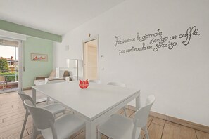 Un espace repas avec une table et des chaises blanches, une pancarte rouge sur laquelle est écrit « Bienvenue », et un mur orné d’une citation inspirante en italien.