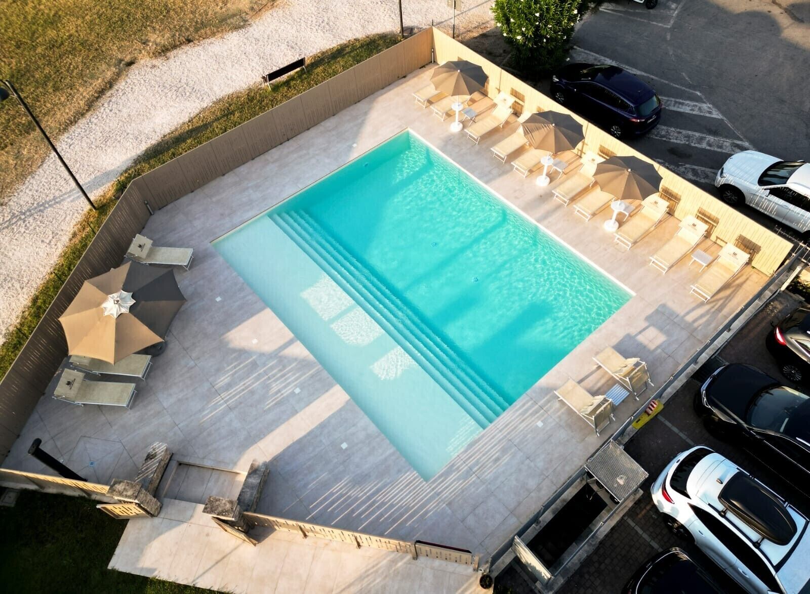 Piscina con sillones de descanso y sombrillas, rodeada de una terraza empedrada y cerca de vehículos estacionados.