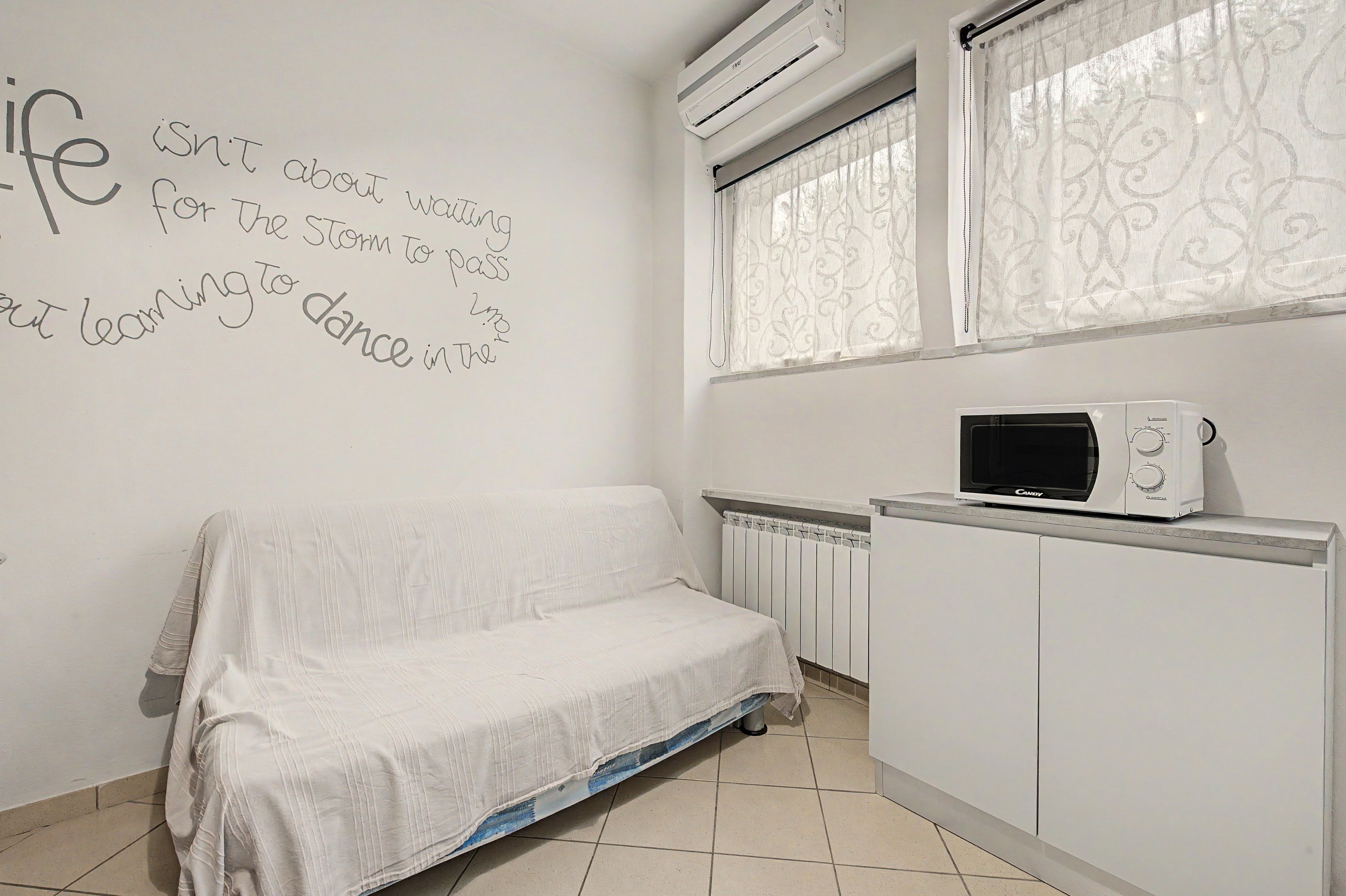 A minimalist room with a white sofa, a wall with motivational writing, a window with patterned curtains, and a microwave on a cabinet.