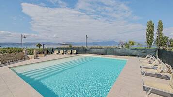 Ein rechteckiger Swimmingpool mit klarem, blauem Wasser, umgeben von einer gefliesten Liegefläche. Es befinden sich Liegestühle und eine überdachte Sitzgelegenheit mit Sonnenschirm am Pool. Im Hintergrund erheben sich ein klarer Himmel und ferne Berge.