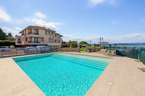 A swimming pool with a clear blue water, surrounded by a tiled deck, a building with balconies, and a view of the sea. - Apartment 'Ibisco - Bilocale Vista Lago' with Lake View, Wi-Fi and Air Conditioning (Sirmione)