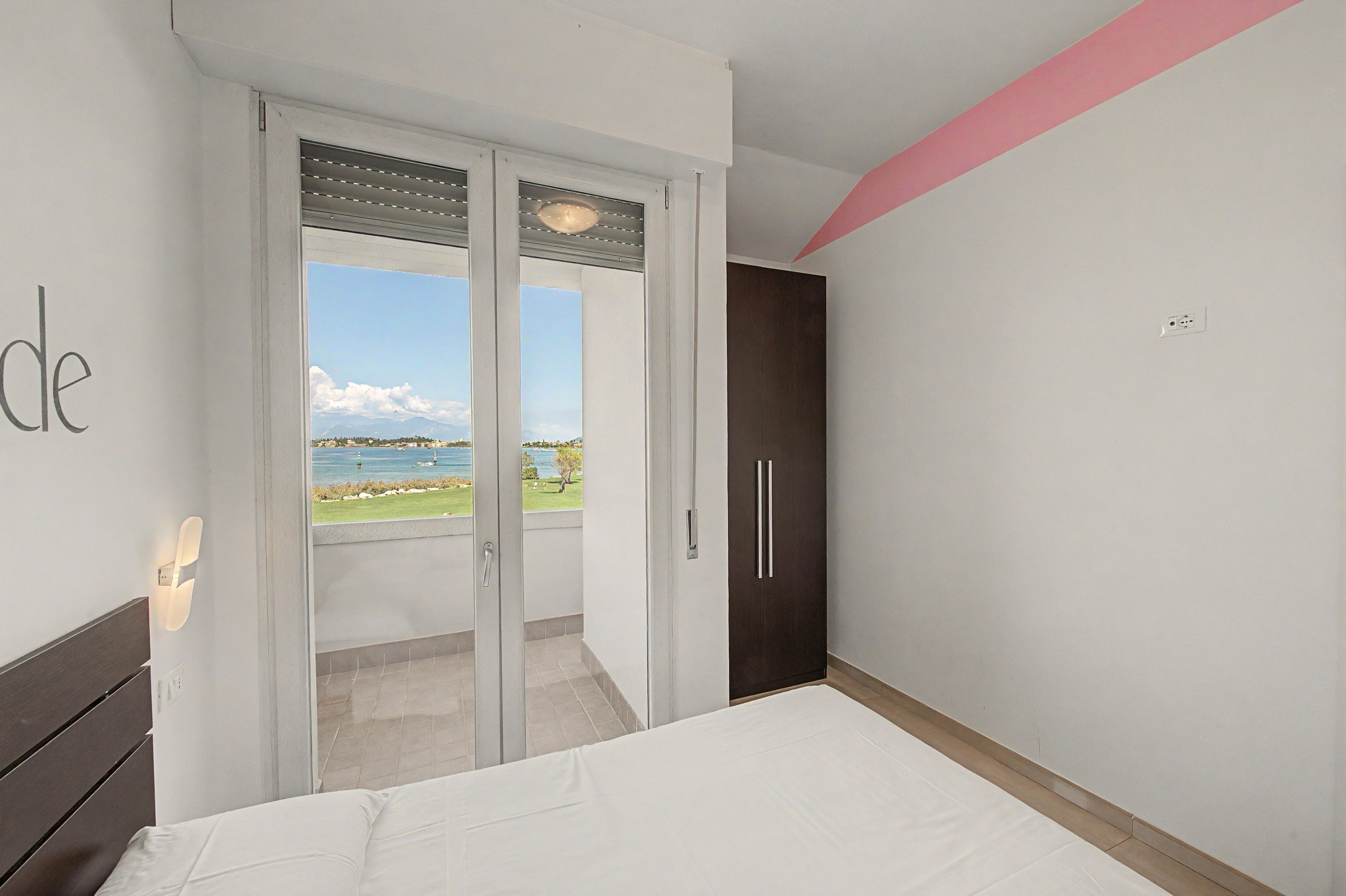 A bedroom with a bed, a wardrobe, and a sliding glass door leading to a balcony with a view of the sea.