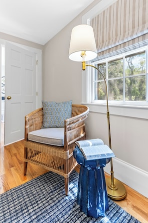 A room with a wicker chair, a blue patterned pillow, a floor lamp, and a window with a view of trees.