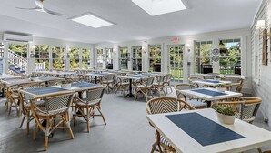 Ampla área de refeições interna, com pé direito alto, grandes janelas e vista para o exterior. As mesas estão dispostas de forma informal, e o espaço possui cadeiras de vime e ventilador de teto.