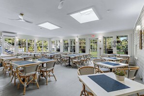 Un comedor interior amplio con techos altos, ventanas grandes y vistas al exterior. Las mesas están dispuestas de forma informal, y la sala cuenta con sillas de mimbre y un ventilador de techo.