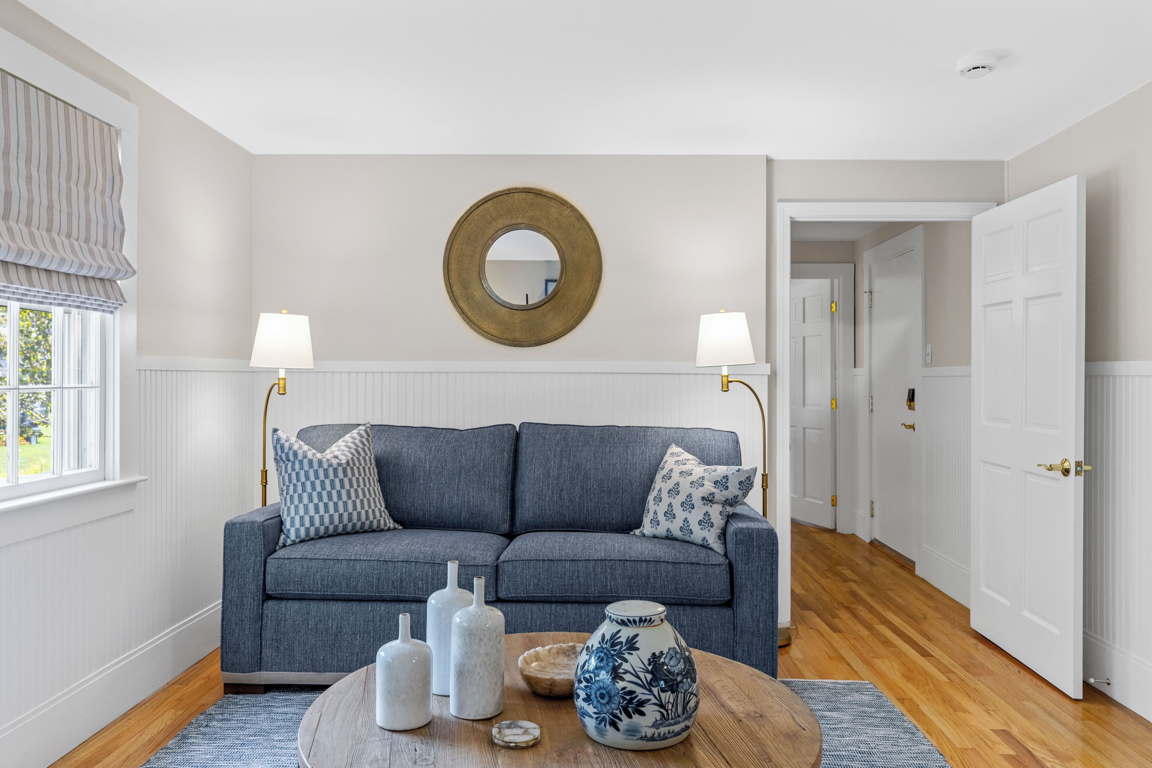 A modern living room with a grey sofa, two lamps, a round mirror, and decorative vases on a wooden coffee table.