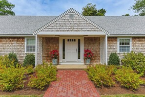 A two-story house with a brick pathway leading to the front door, flanked by two potted plants with red flowers.