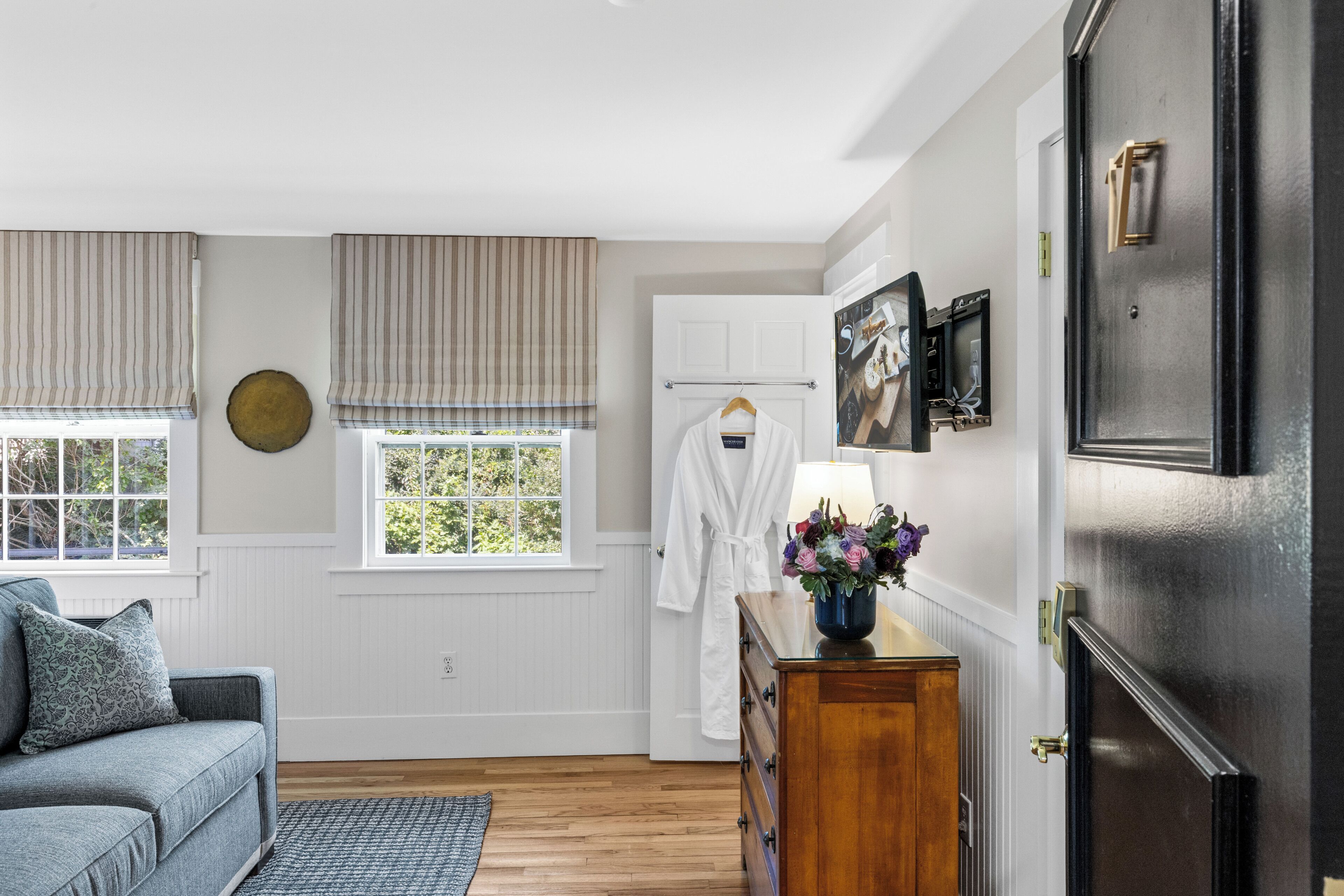 A room with a wooden dresser, a white robe, a window with blinds, and a wall-mounted picture.