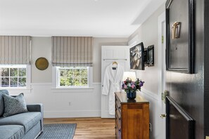 A room with a wooden dresser, a white robe, a window with blinds, and a wall-mounted picture.