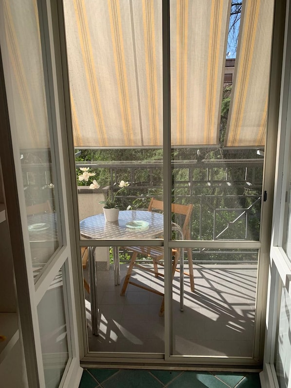 Puerta corrediza de vidrio que da a un balcón exterior con una mesa redonda y sillas, una planta en maceta y vistas de un edificio con balcón.