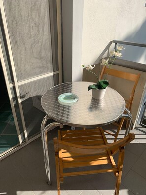 Een ronde metalen tafel met een glazen blad, gecombineerd met een houten stoel, op een balkon met uitzicht op een gebouw.