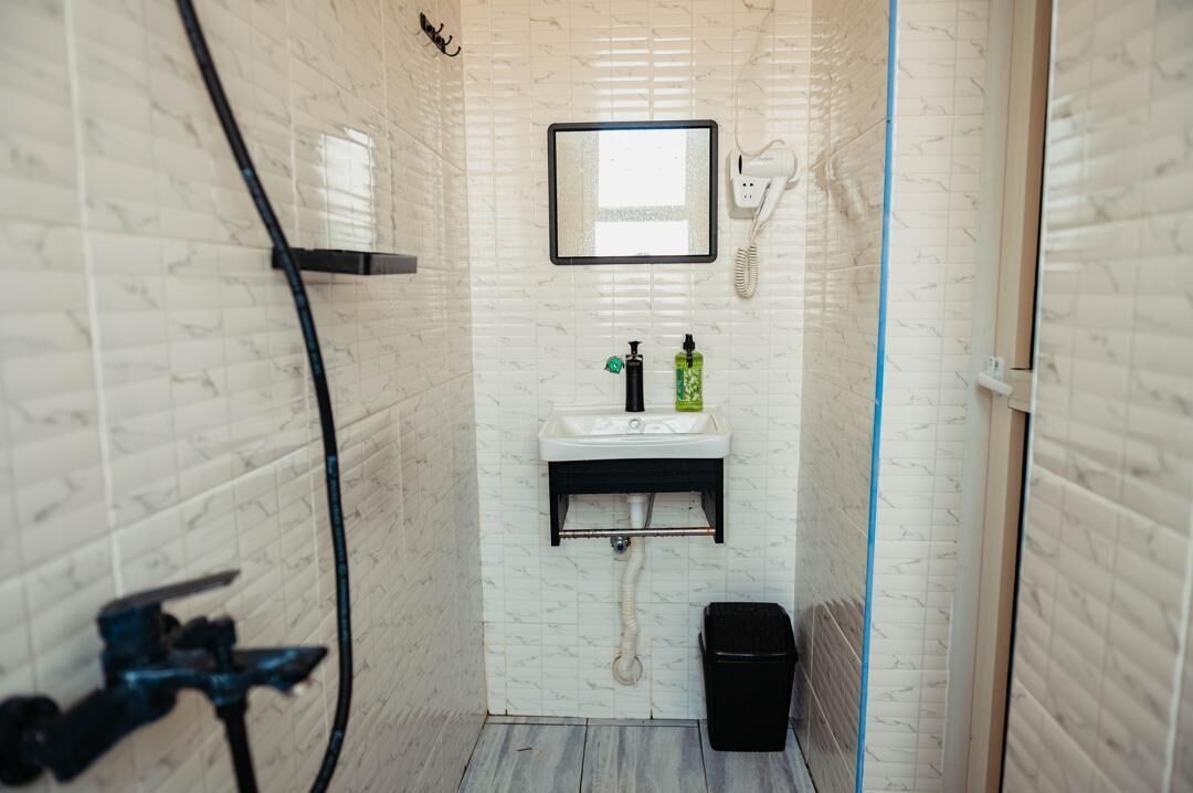 Casa de banho com parede revestida a azulejos brancos, uma pia com espelho em cima, secador de cabelo e um contentor de lixo preto.