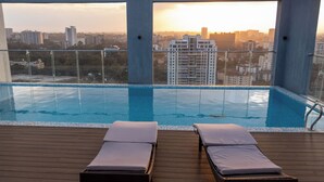 Une piscine à débordement avec deux chaises longues, entourée d’une terrasse en bois, offrant une vue sur le paysage urbain au coucher du soleil.