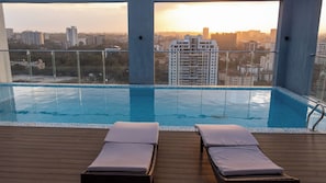 Ein Infinity-Pool mit zwei Liegestühlen, umgeben von einer Holzterrasse, mit Blick auf eine Skyline bei Sonnenuntergang.
