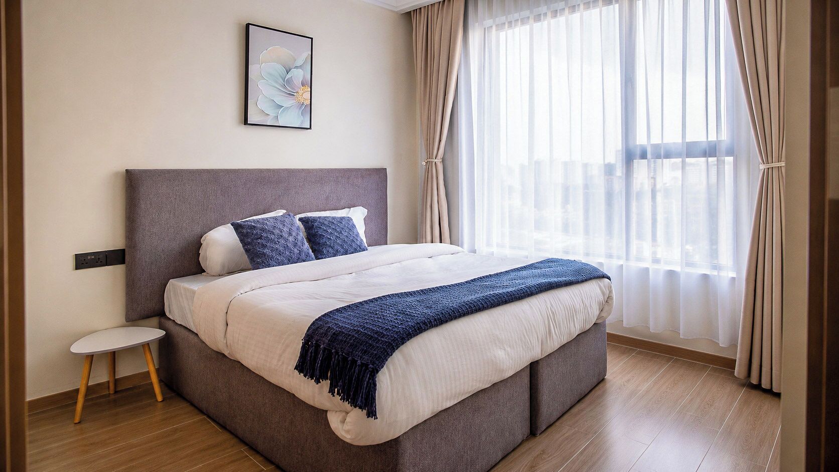 A modern bedroom with a large bed, a small bedside table, and a city view through sheer curtains.