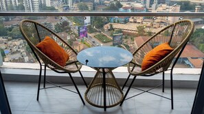 Ein Balkon mit zwei Korbstühlen und einem kleinen runden Tisch, mit Blick auf eine Stadtlandschaft mit Gebäuden und einer Straße.