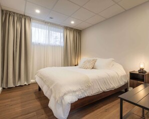 Un dormitorio con una cama grande, suelo de madera, una mesita de noche con lámpara y un ventanal con cortinas.