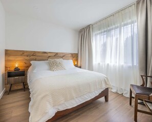 A bedroom with a wooden headboard, a bed with white bedding, a nightstand with a lamp, a chair, and a window with curtains.