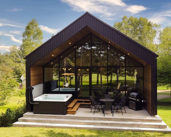 A modern outdoor hot tub area with a wooden bench and chairs, surrounded by glass walls.
