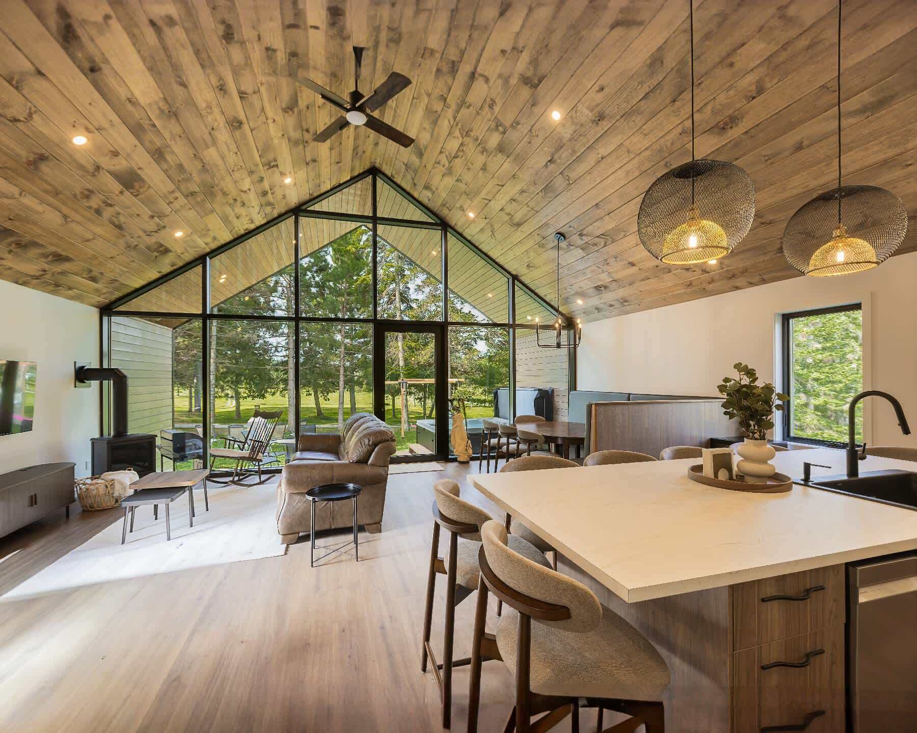 A spacious kitchen with a large island, wooden ceiling, and large windows offering a view of the outdoors.