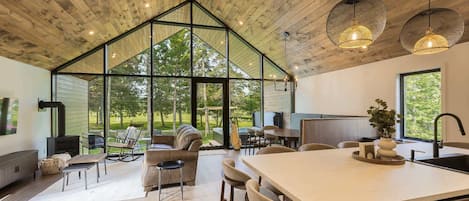 A spacious kitchen with a large island, wooden ceiling, and large windows offering a view of the outdoors.