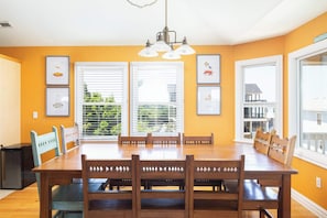 Uma sala de jantar com mesa e cadeiras de madeira, janelas amplas com persianas e quadros nas paredes.