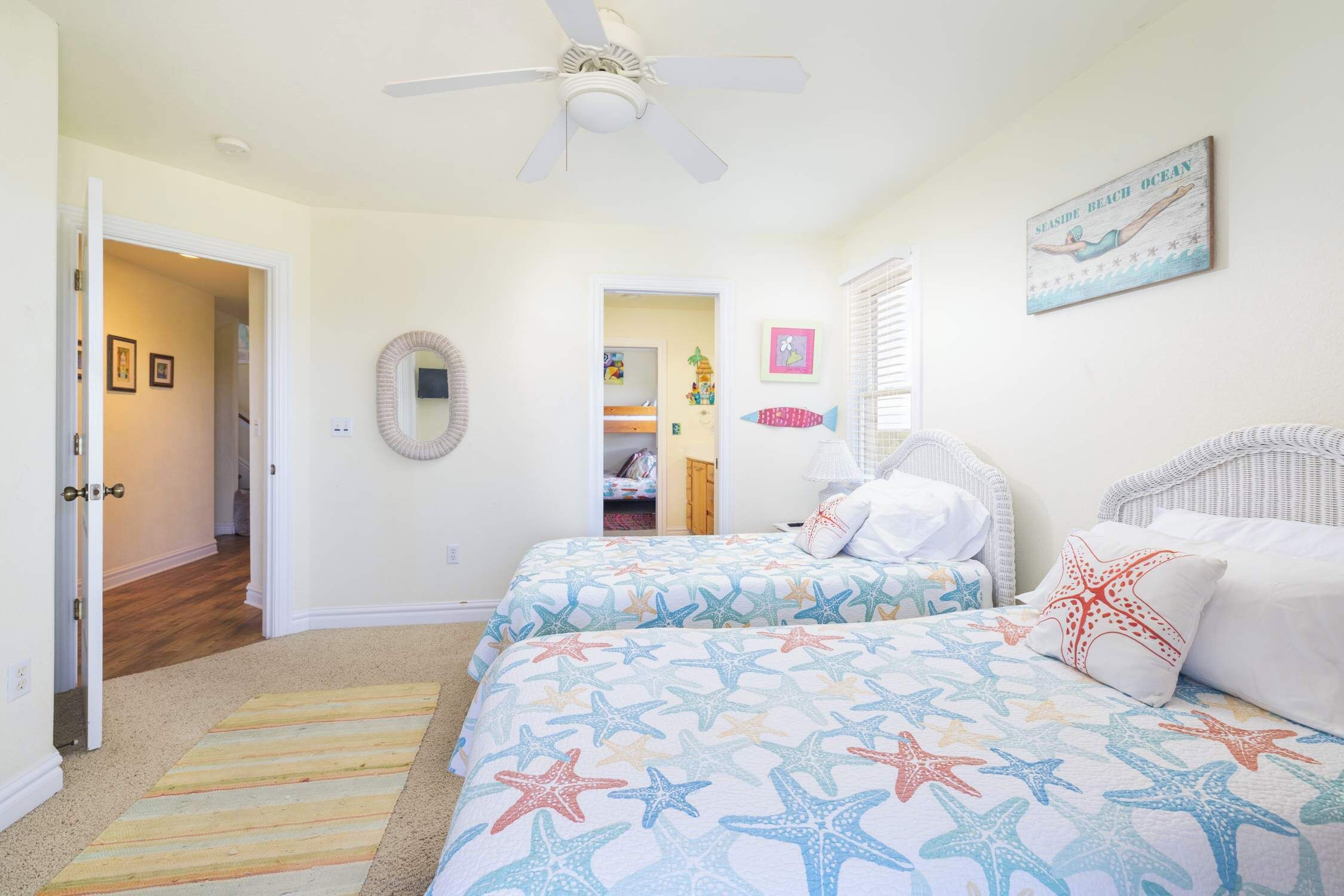 Une chambre avec deux lits, un ventilateur de plafond, un tableau sur le thème de la plage et un couloir menant à une autre pièce.