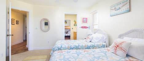 Une chambre avec deux lits, un ventilateur de plafond, un tableau sur le thème de la plage et un couloir menant à une autre pièce.