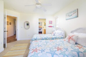 A bedroom with two beds, a ceiling fan, a beach-themed painting, and a hallway leading to another room. - 2 min Walk to Beach w Ocean Views, Pool, Game Room (Nags Head)