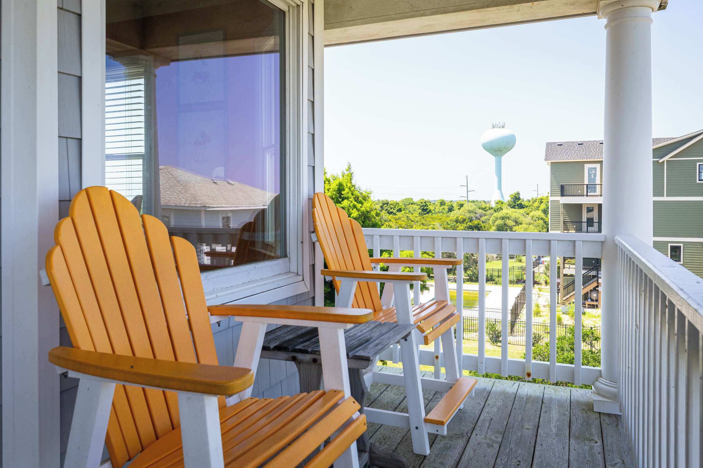 A porch with wooden chairs and a table, overlooking a residential area with houses and a water tower.
