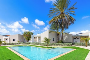 A swimming pool with a clear blue sky, palm trees, and modern buildings. - Casa Atardi – Your tropical home away from home in Curaçao (Willemstad)