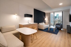 Ein modernes Hotelzimmer mit einer Küchenzeile, einer Sitzecke mit Sofa und Sesseln, einem Esstisch und Blick auf die Stadt.