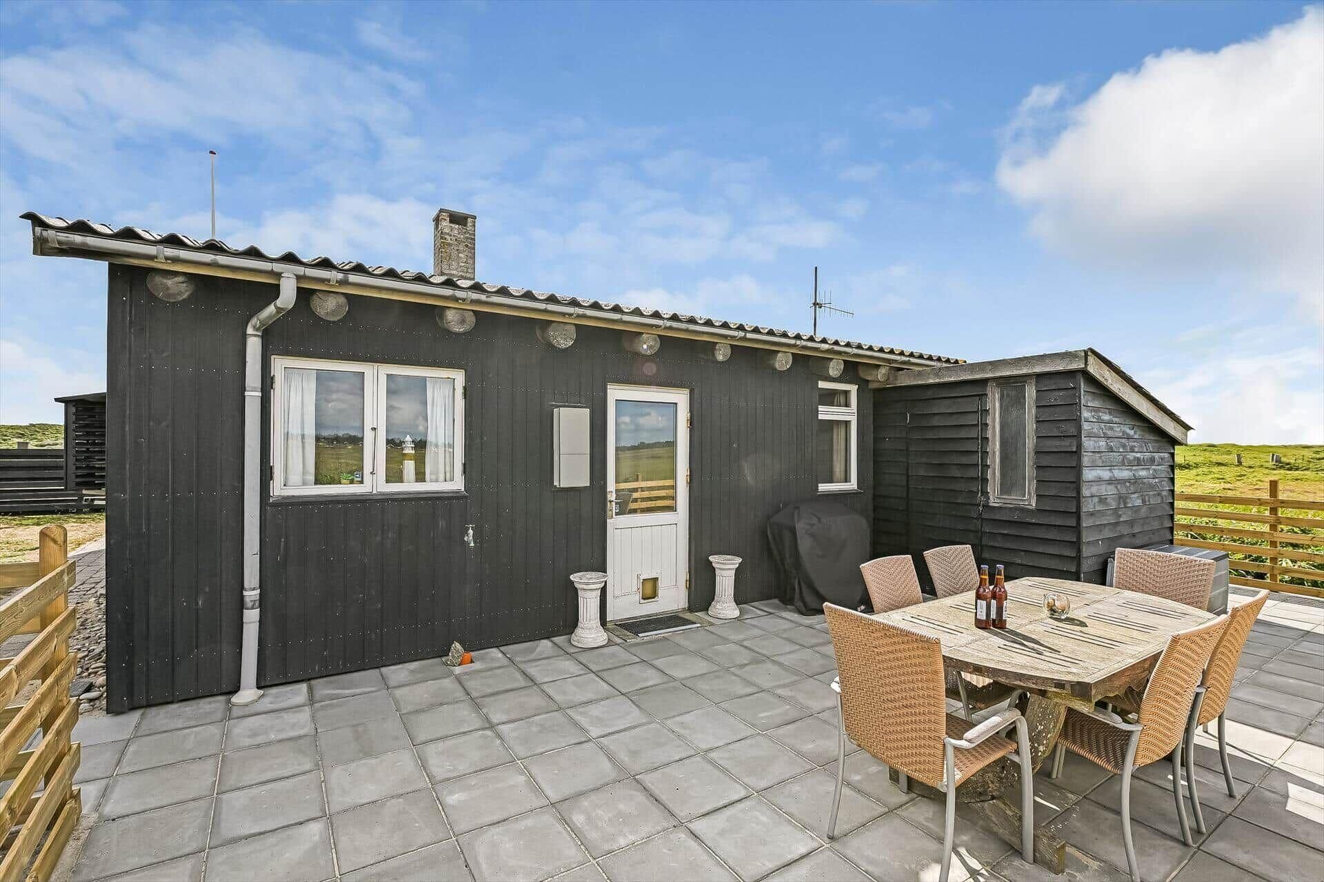 Une petite maison en bois sombre, dotée d’une terrasse carrelée, d’une table avec des chaises et offrant une vue sur la campagne.