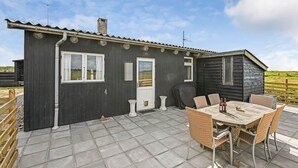 Een klein, donker houten huisje met een betegeld terras, een tafel met stoelen en uitzicht op het platteland.