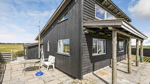Een houten huis met twee verdiepingen, een overdekte veranda, een klein terras met tafel en stoel, en uitzicht op het omliggende landschap.