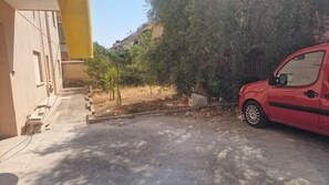A red car parked in a driveway with a building on the left and a tree in the background.