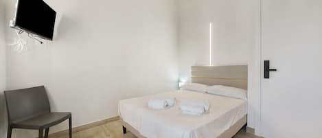 A hotel room with a bed, a chair, a flat-screen TV mounted on the wall, and a door.