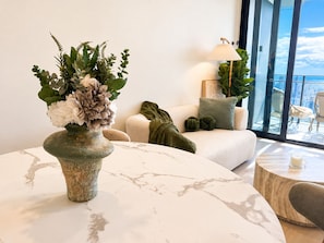 A modern living room with a marble table, a vase with flowers, a sofa, and a view of the city through large windows. - Sage Above the Sea - Ocean Views (Broadbeach)