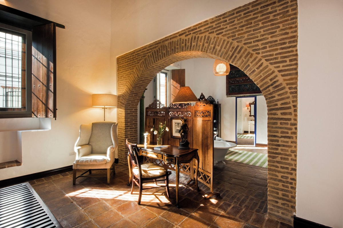 Een kamer met een bakstenen boog, een stoel, een tafel, een lamp en uitzicht op een badkamer.