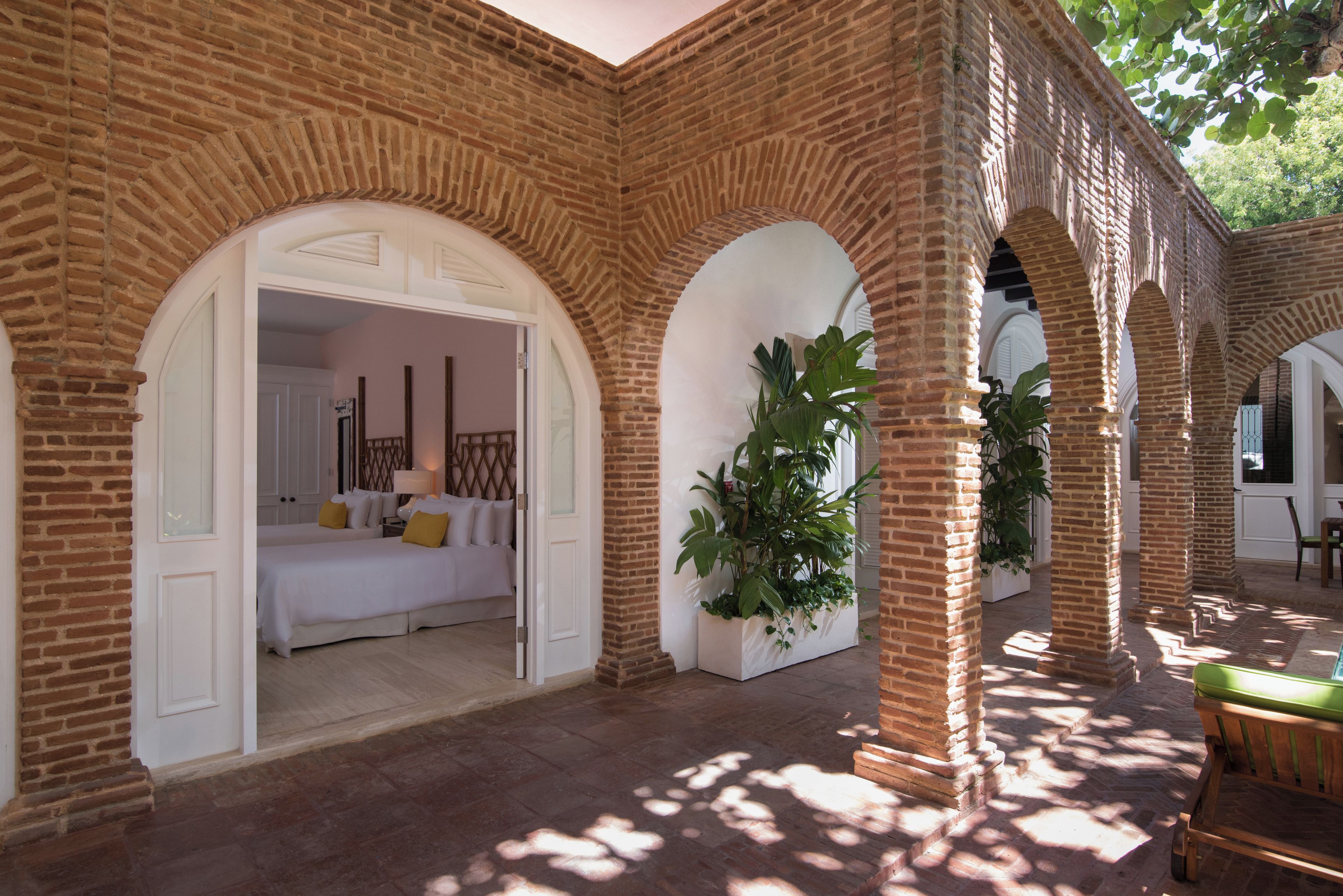 A hotel room with a brick archway leading to a bed, a potted plant, and a view of the outdoors.