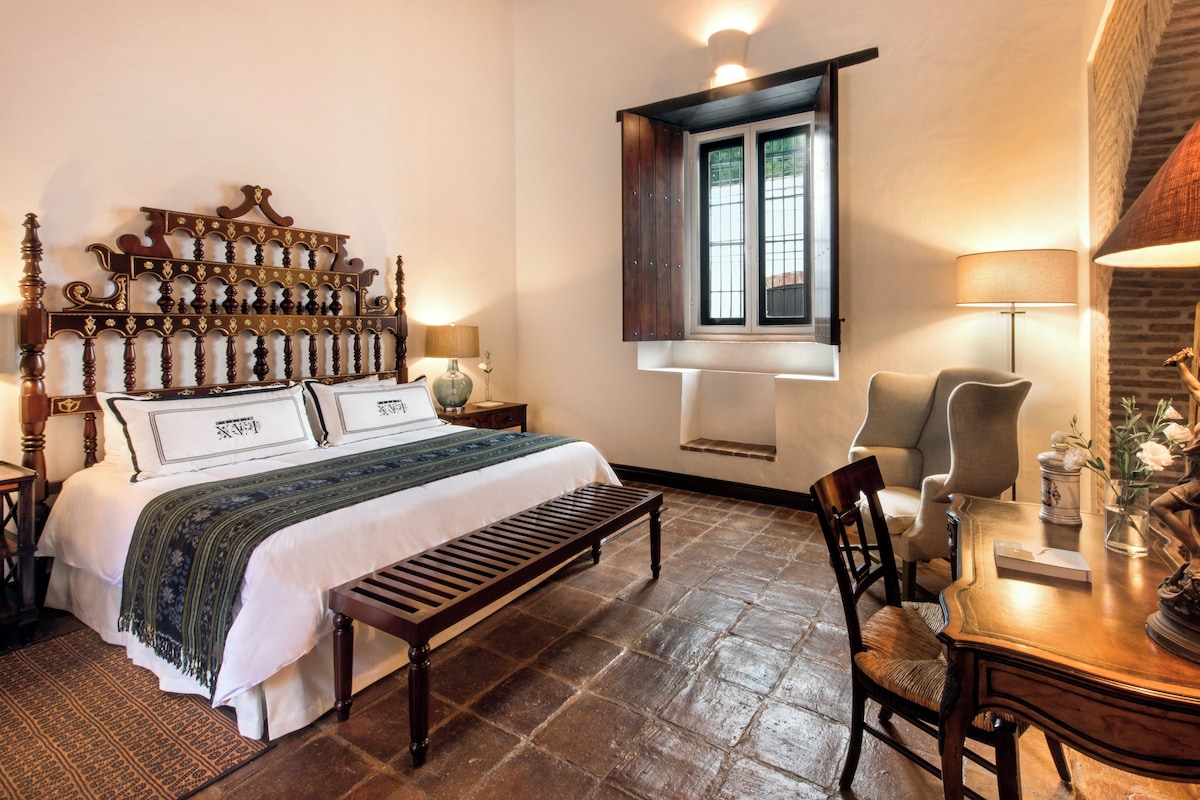 A traditional bedroom with a wooden four-poster bed, a bench, a desk, and a statue.