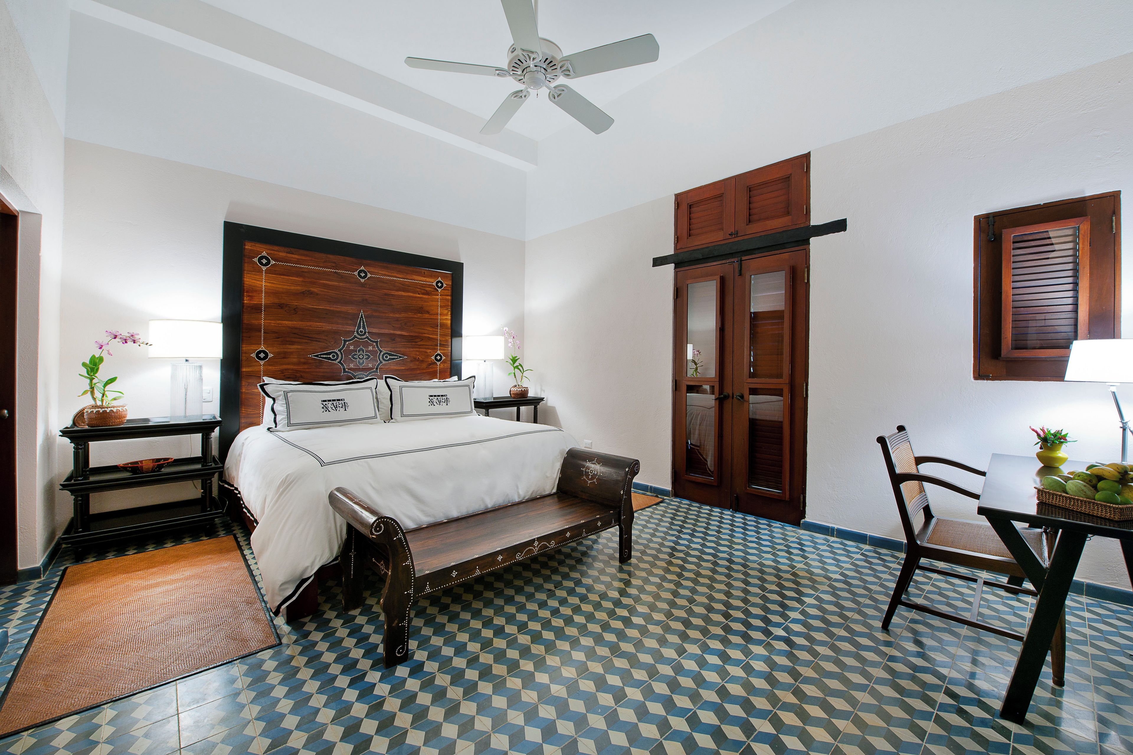 Quarto com cama, um banco de madeira, mesa com cadeira, ventilador de teto e janela com persianas.