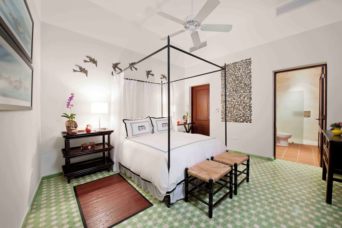 A bedroom with a four-poster bed, a ceiling fan, a decorative wall piece, and a view of the bathroom.