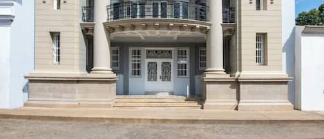 Un bâtiment de style classique, avec une entrée centrale flanquée de colonnes, un balcon orné de balustrades et un ciel dégagé au-dessus.