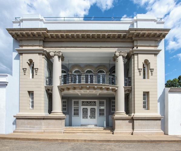 A classical building with a central entrance flanked by columns, a balcony with ornate railings, and a clear sky above.