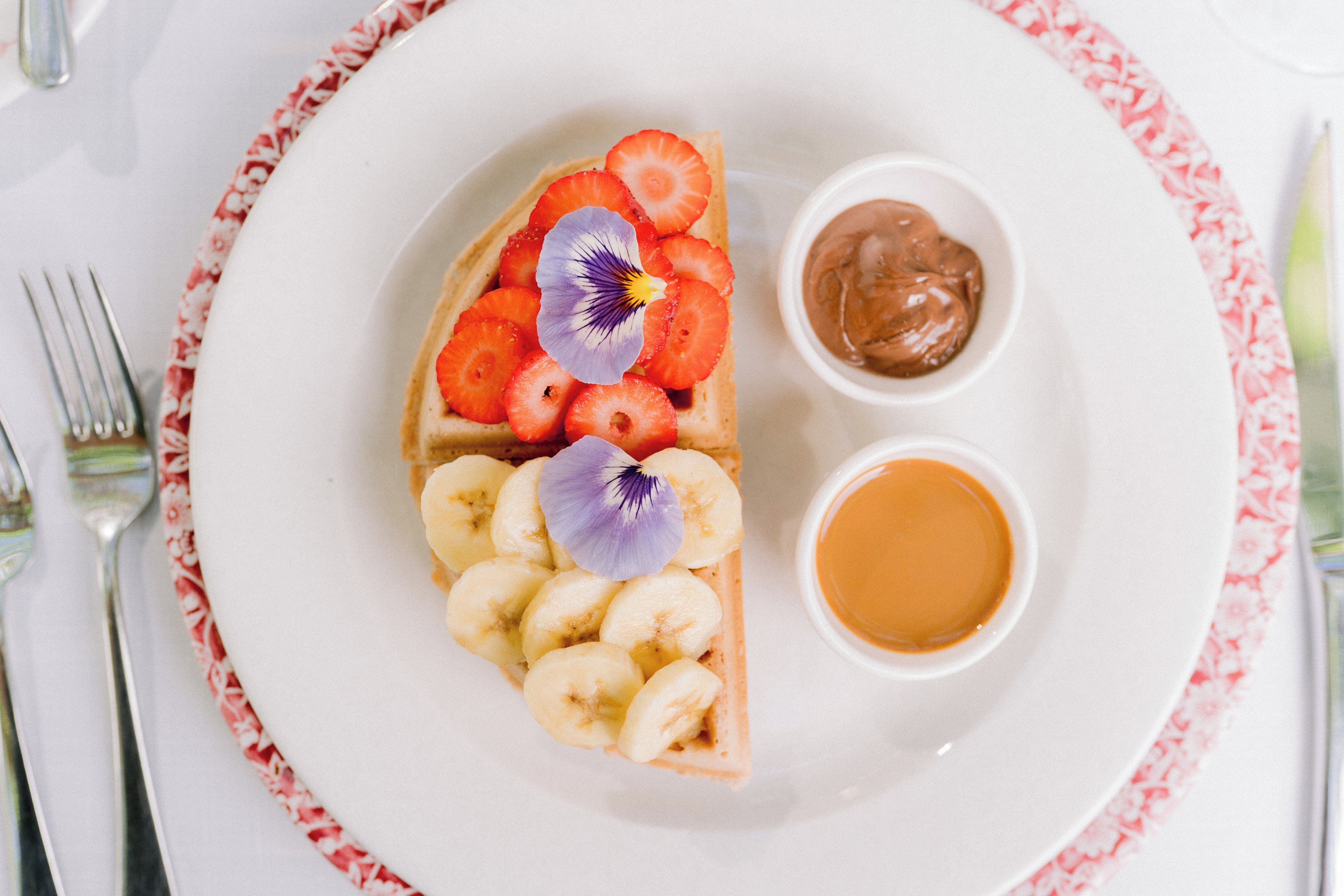 A plate with a slice of pizza topped with tomatoes, basil, and a flower, accompanied by a small bowl of sauce and a cup of tea.