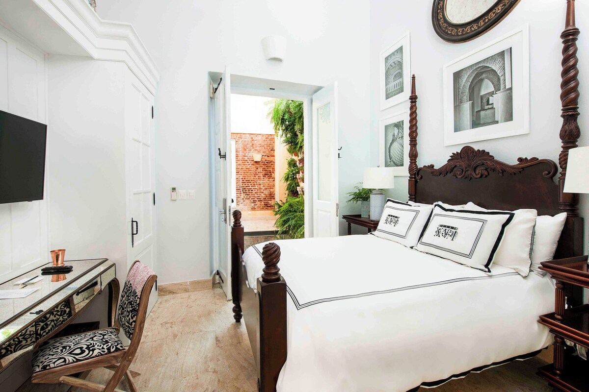 A hotel room with a bed, a desk with a computer, a chair, a television, and a door leading to a courtyard.