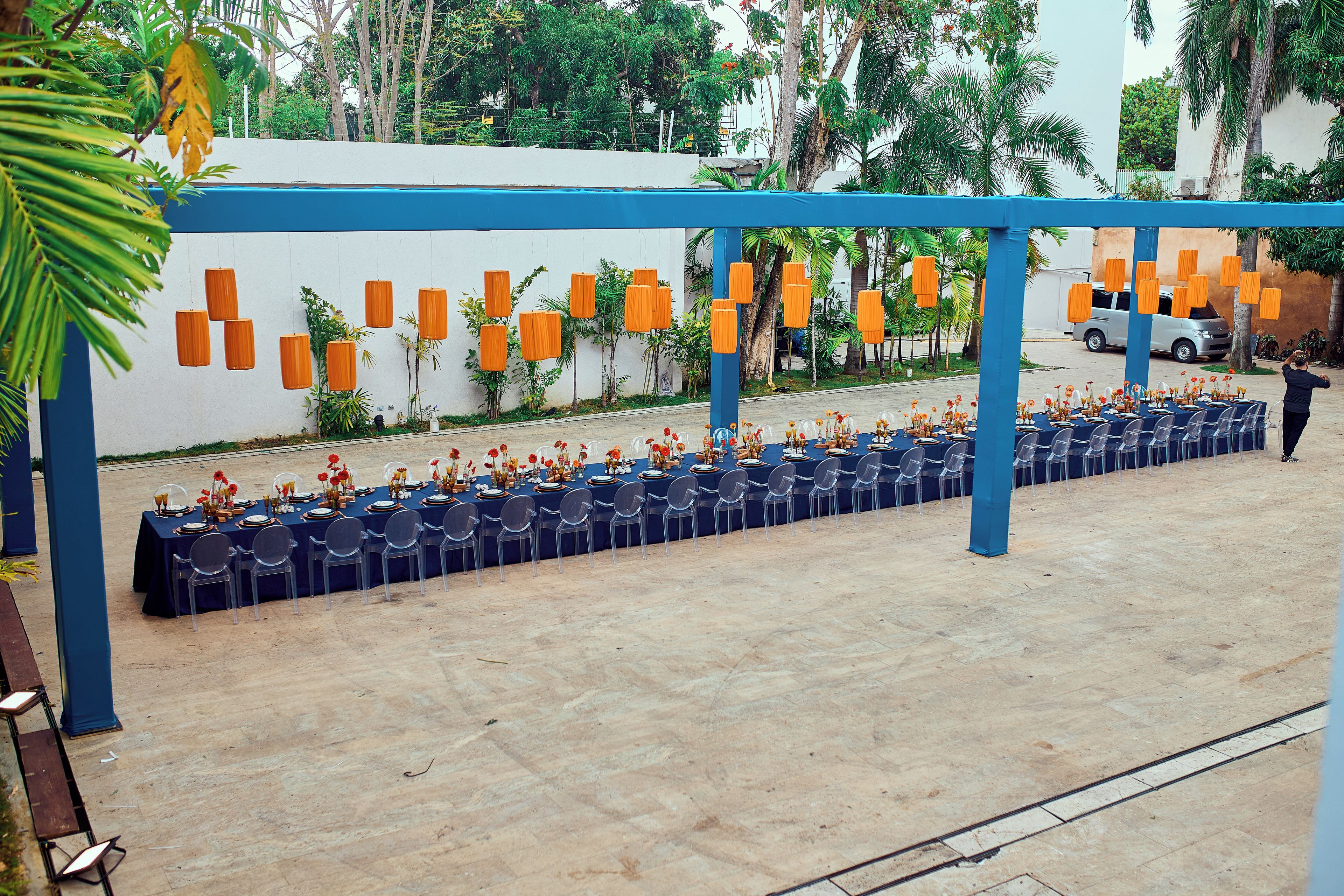 An outdoor dining area with tables set for guests, a blue canopy overhead, and a white wall with hanging orange lanterns.