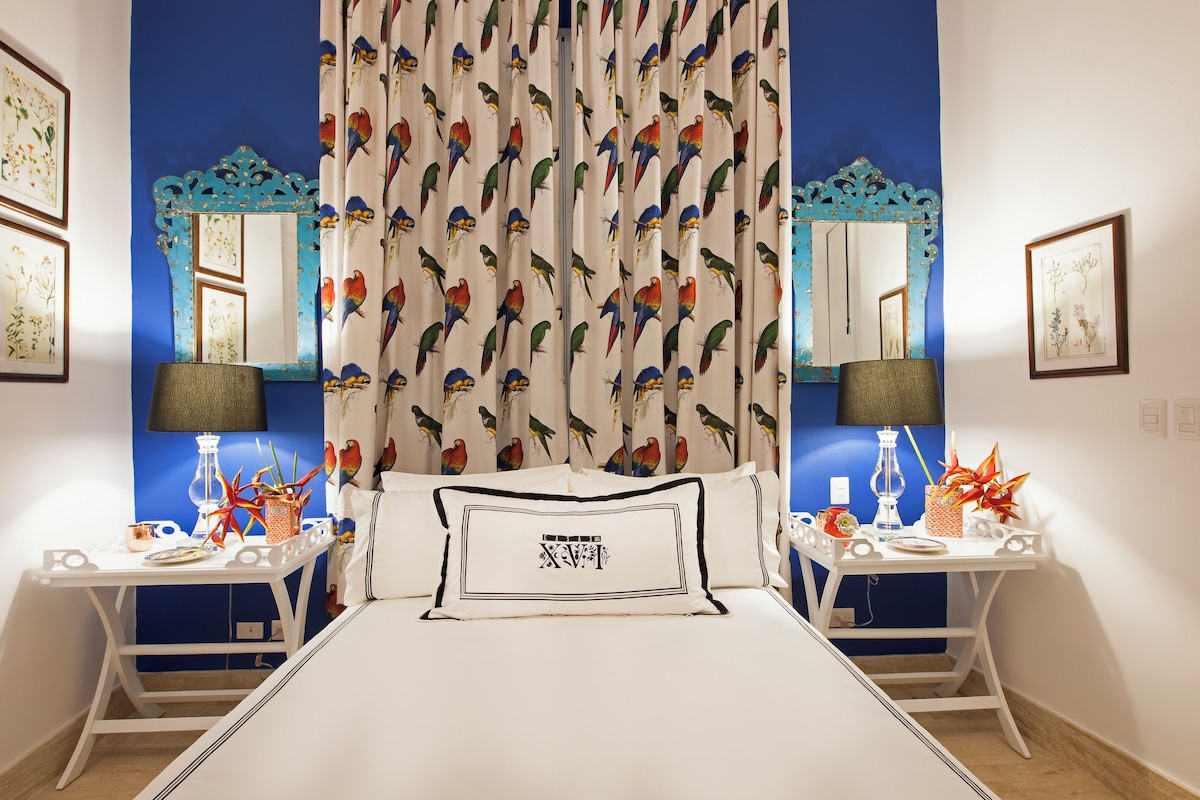 Een slaapkamer met een bed, twee nachtkastjes, lampen, een spiegel en gordijnen met een papegaaienprint.
