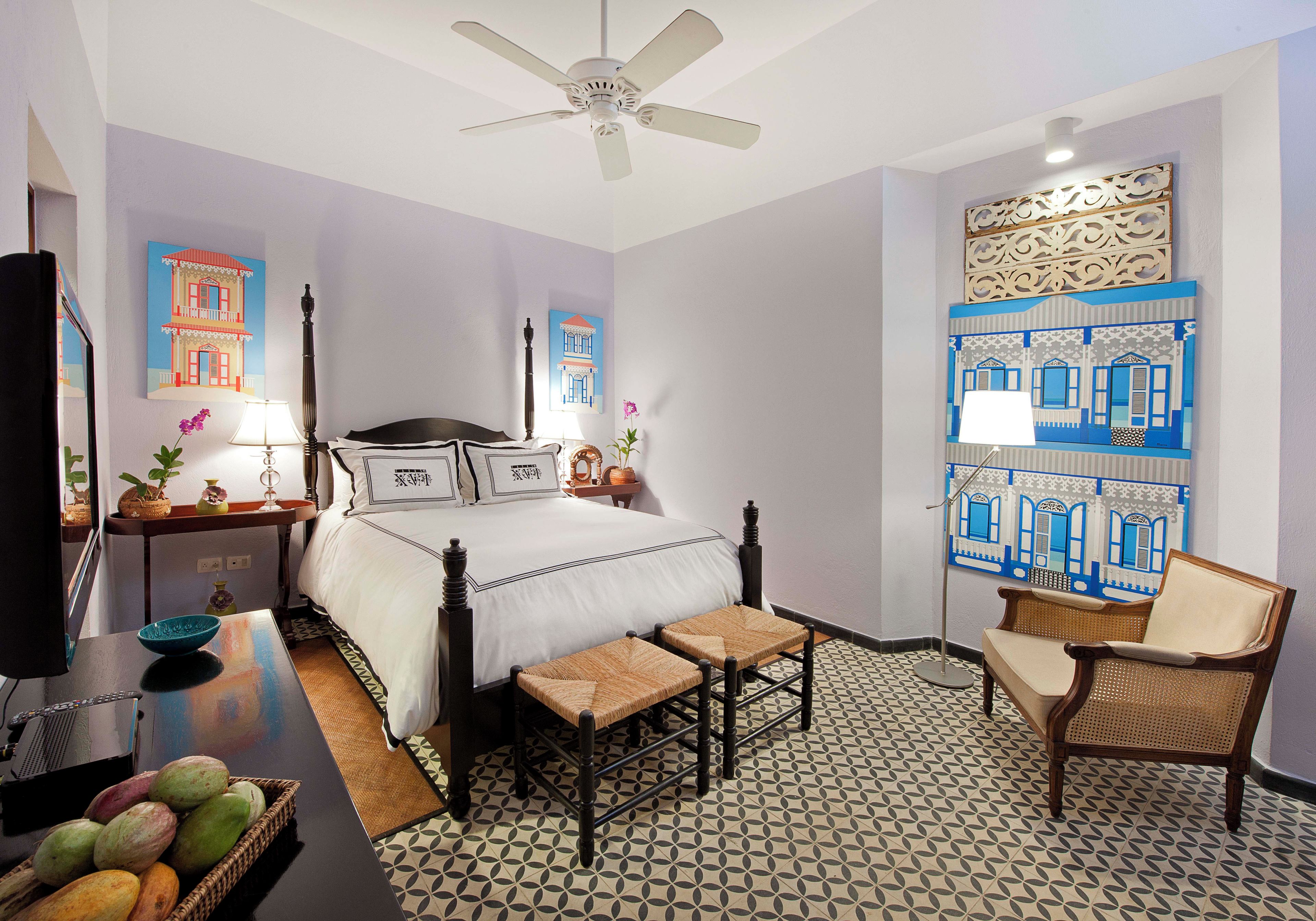 Une chambre d’hôtel avec un lit, une chaise, une table, un ventilateur de plafond et des œuvres d’art accrochées aux murs.
